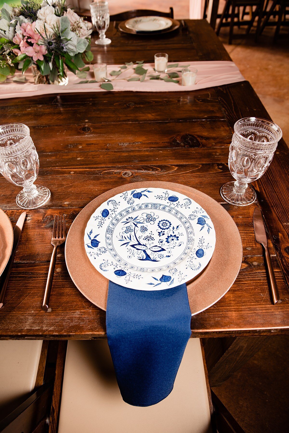 Dark wood farm table set with a gold charger, clear crystal goblet, a blue linen napkin and a blue vintage china plate. Gold flatware is set on either side of the vintage plate. The center of the table has a blush sheer table runner and a low floral centerpiece with blush and white flowers with eucalyptus.