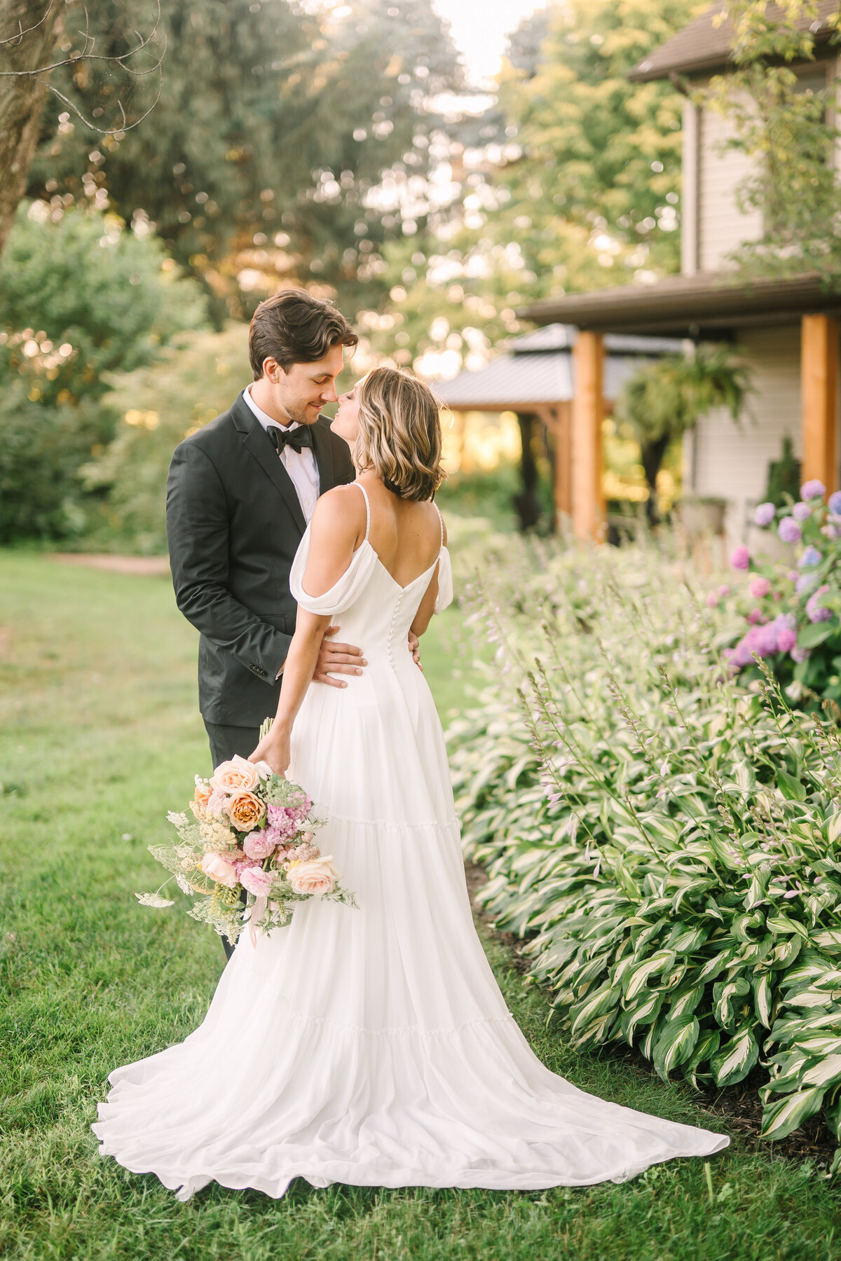 a bride and groom posing in the front yard gardens of the modern farmhouse overnight accommodations at Willowbrook wedding venue with a blush bridal bouquet of fresh flowers