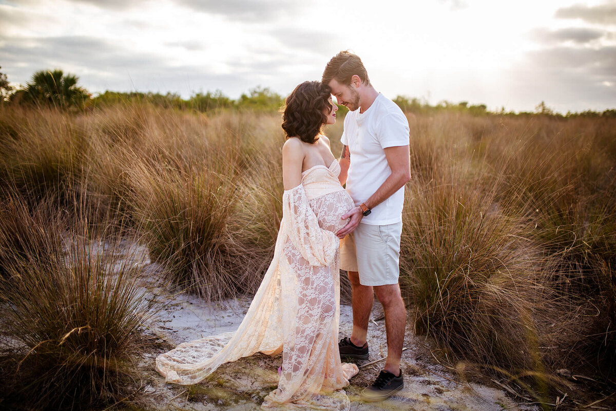 Woman and man cradling pregnant belly standing in tall seagrass during golden light hour at Cypress Point Park in Tampa, FL 