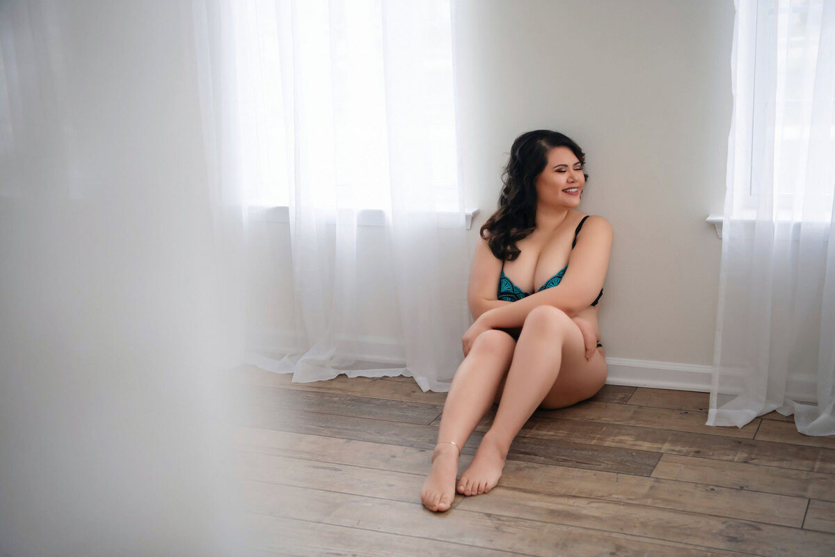 Woman in teal lingerie sits on wooden floor by white curtains, smiling confidently. The scene is softly lit, creating a calm and intimate mood.
