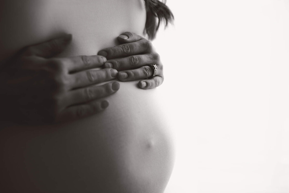 Brittany Danielle captured this up close black and white Maternity portrait. A  woman puts two hands on top of her belly. She is wearing a white shirt and there is a bright window light in behind her. Photo by Brittany Danielle