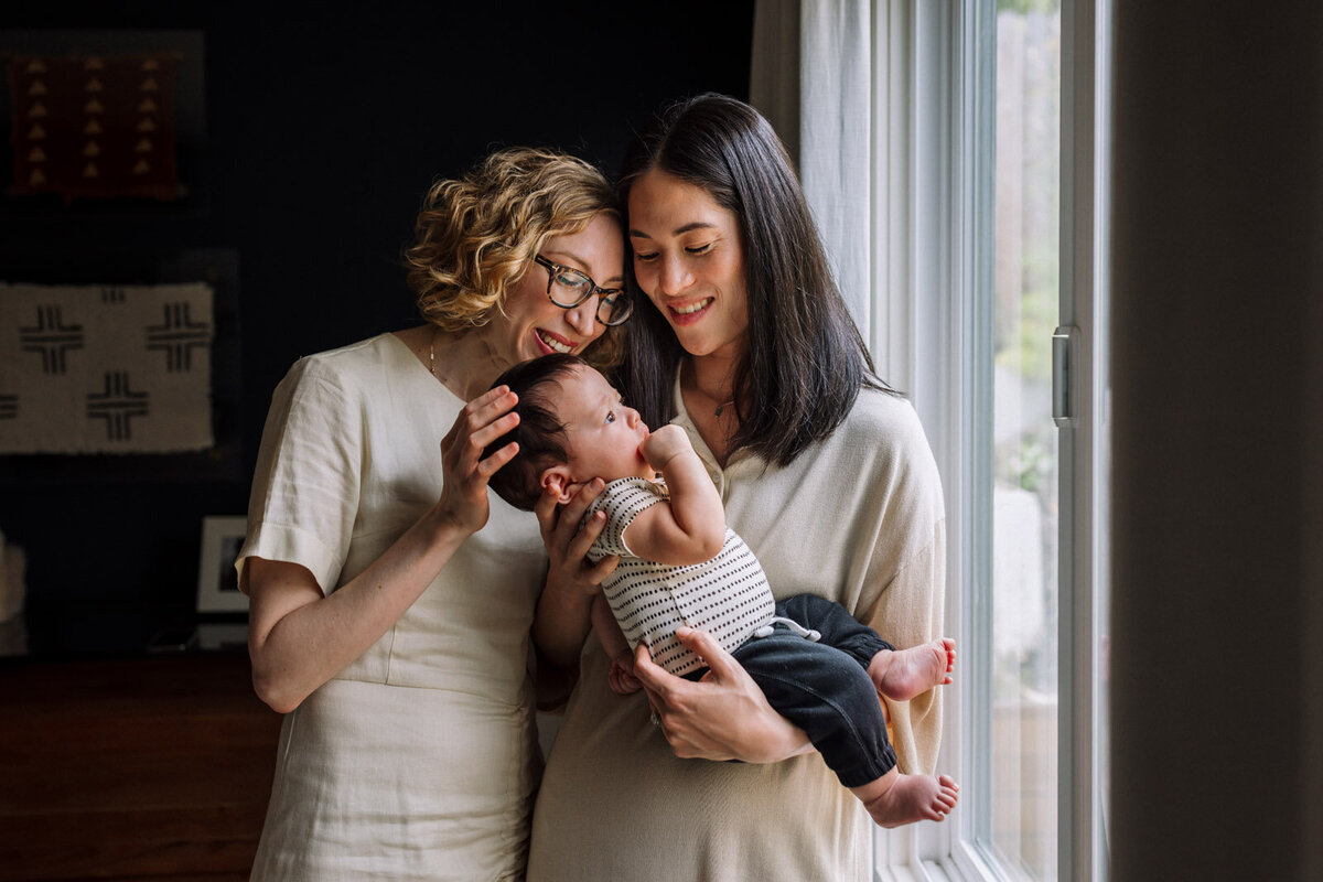 two moms in off white holding baby by window and smiling at him