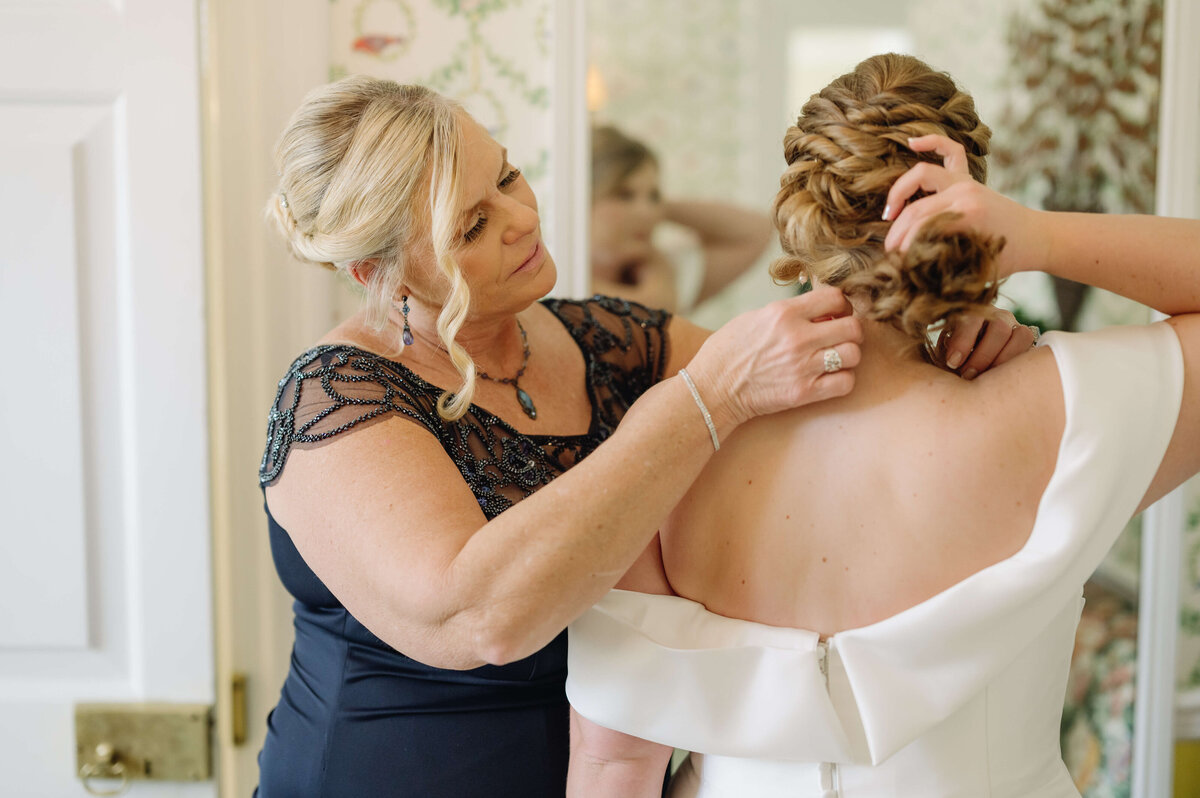 mother of the bride helps bride put on her wedding jewelry in the bridal suite captured by Virginia wedding photographer before the wedding day begins