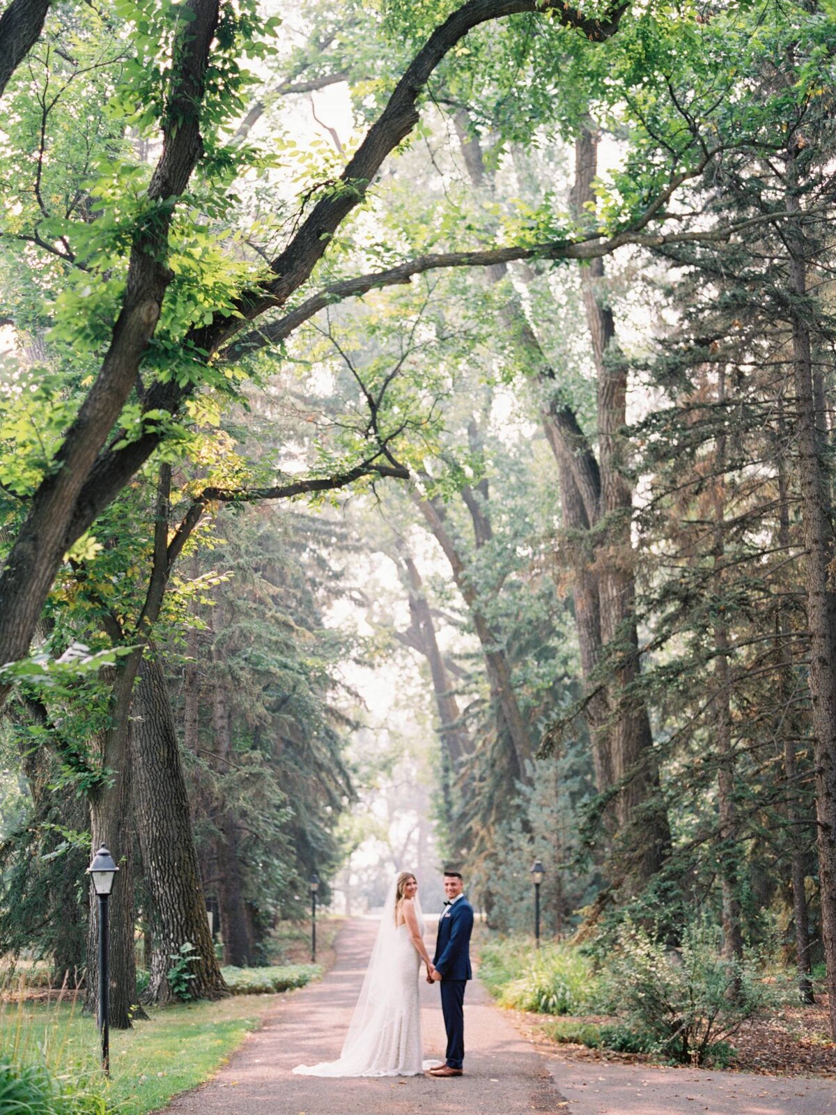 Bride and groom on the tree lined path at The Norland Historic Estate, a classic vintage wedding venue in Lethbridge, AB, featured on the Brontë Bride Vendor Guide.