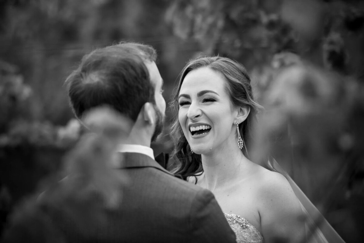A joyous black and white moment captured at a wedding, featuring a bride laughing heartily as she looks at her groom, with soft focus on the surrounding foliage.
