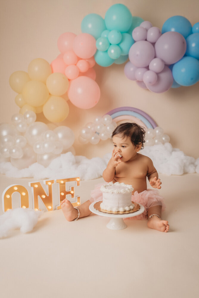 Photo shoot at Atlanta, Georgia cake smash photographer studio with one year old little girl in pink tutu smashing a cake with rainbow pastel balloon garland behind her with white fluffy clouds.