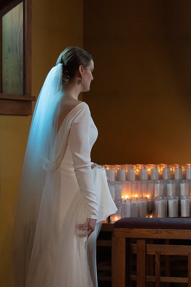 Bride in open-backed gown with long veil enters candled vestibule during wedding ceremony
