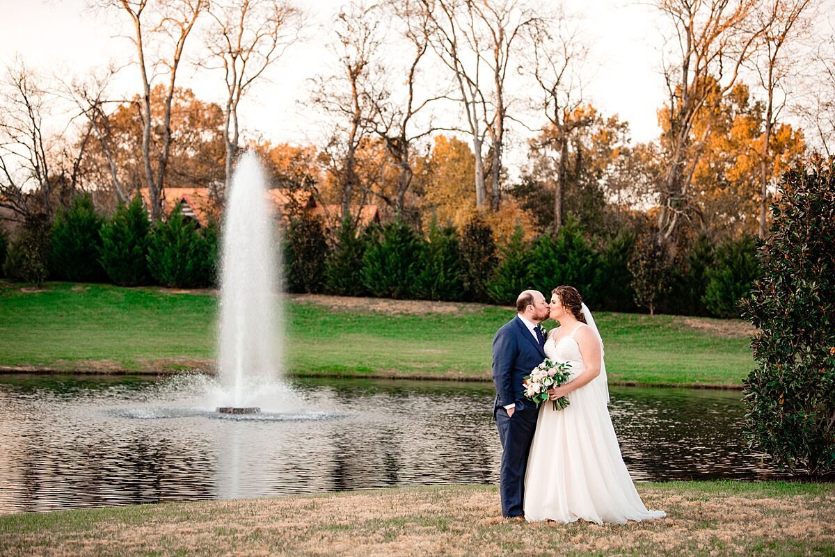 Fall wedding at Sycamore Farms with bride and groom kissing in front of a large fountain in a pond at Sycamore Farms.