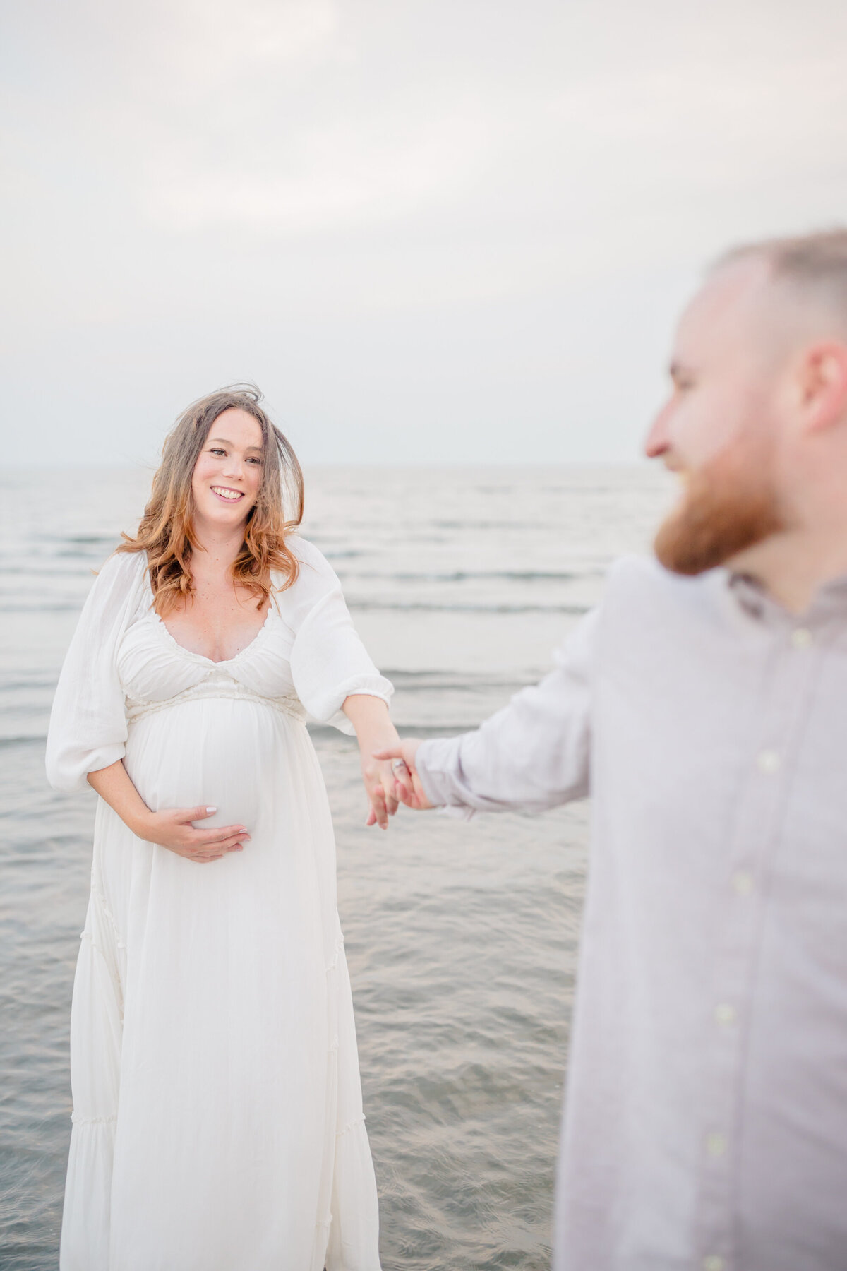 A pregnant woman in a flowing white dress stands in the shallow ocean water, smiling as she holds hands with her partner