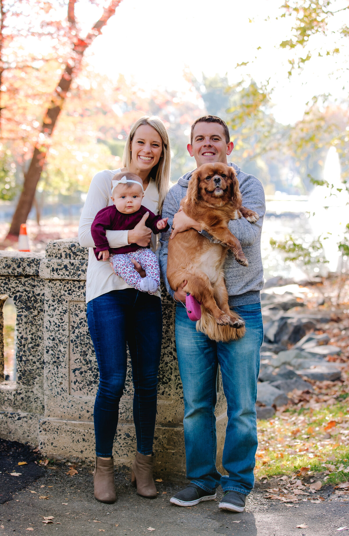 parents holding baby girl and dog in park in front of autumn trees
