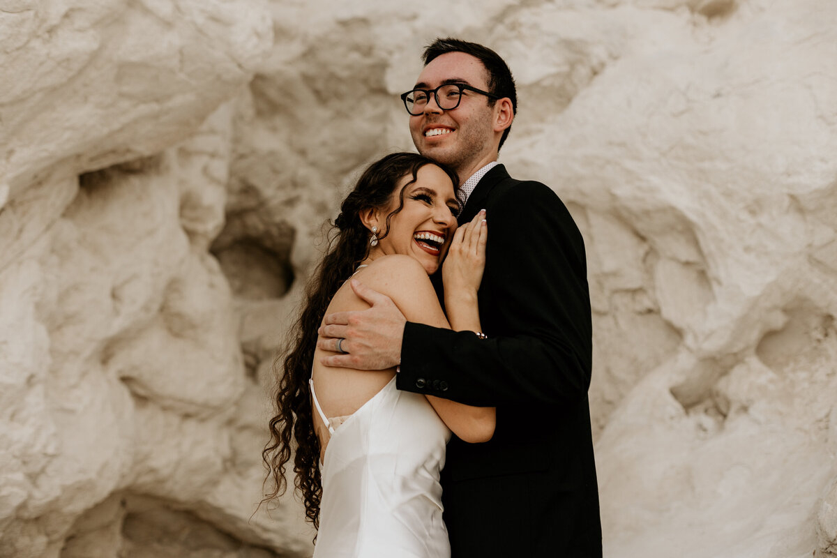 bride and groom hugging and looking off in front of a white rock