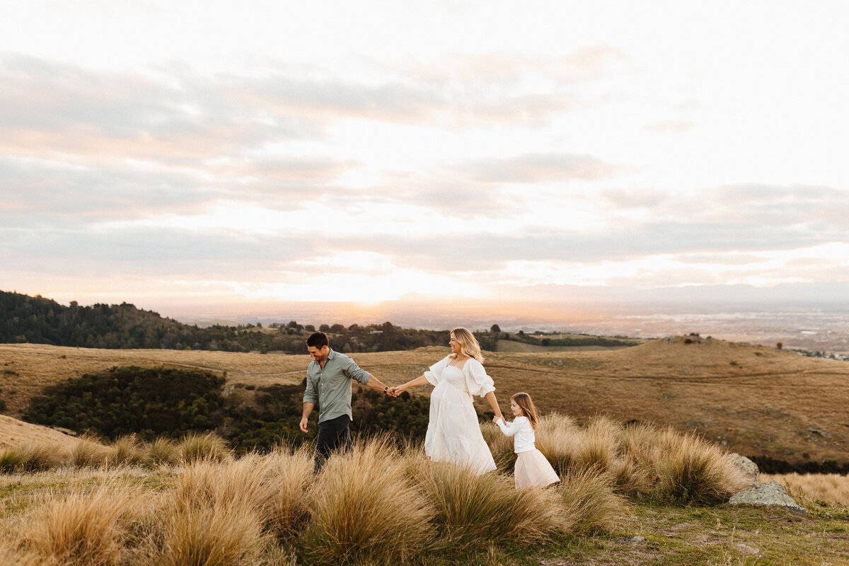 A family of three walking along a paddock of long grasses during a maternity family photoshoot in christchurch with family photographer lucy jane photography