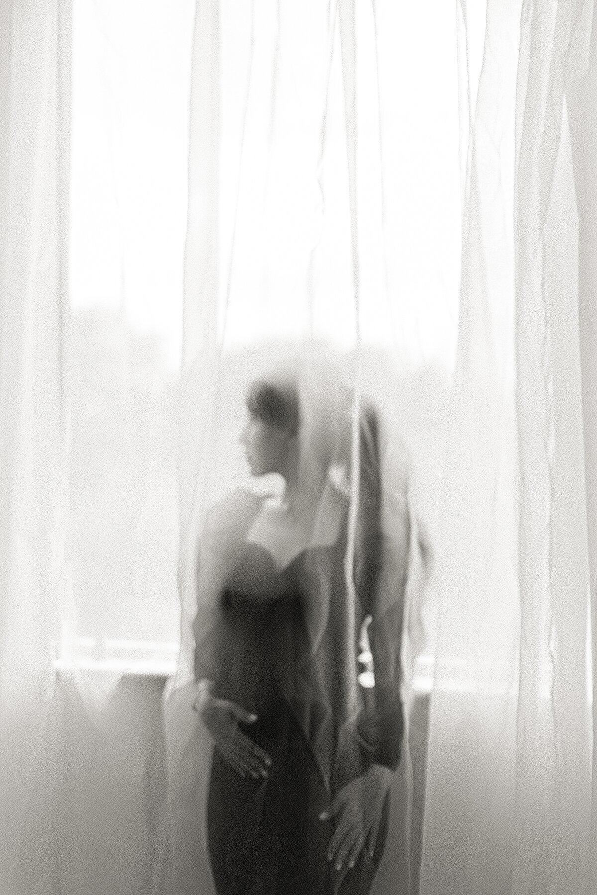 Soft out of focus black and white photo of a woman dressed in all back behind a thin layer if sheer drapes as she looks over her shoulder and out of the window in a Dallas photography studio.