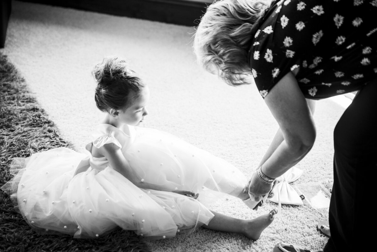 A small flower girl sits on the ground in her fluffy dress as a woman helps her put on her shoes.
