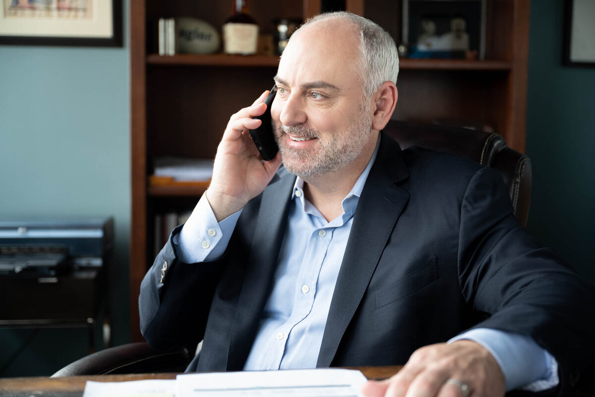A portrait of a man sitting behind a desk talking on his cellphone looking to the window