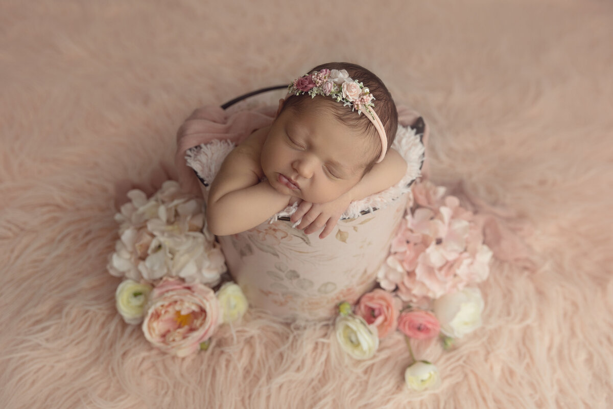 A newborn baby girl sleeps inside a floral print bucket wearing a pink floral headband