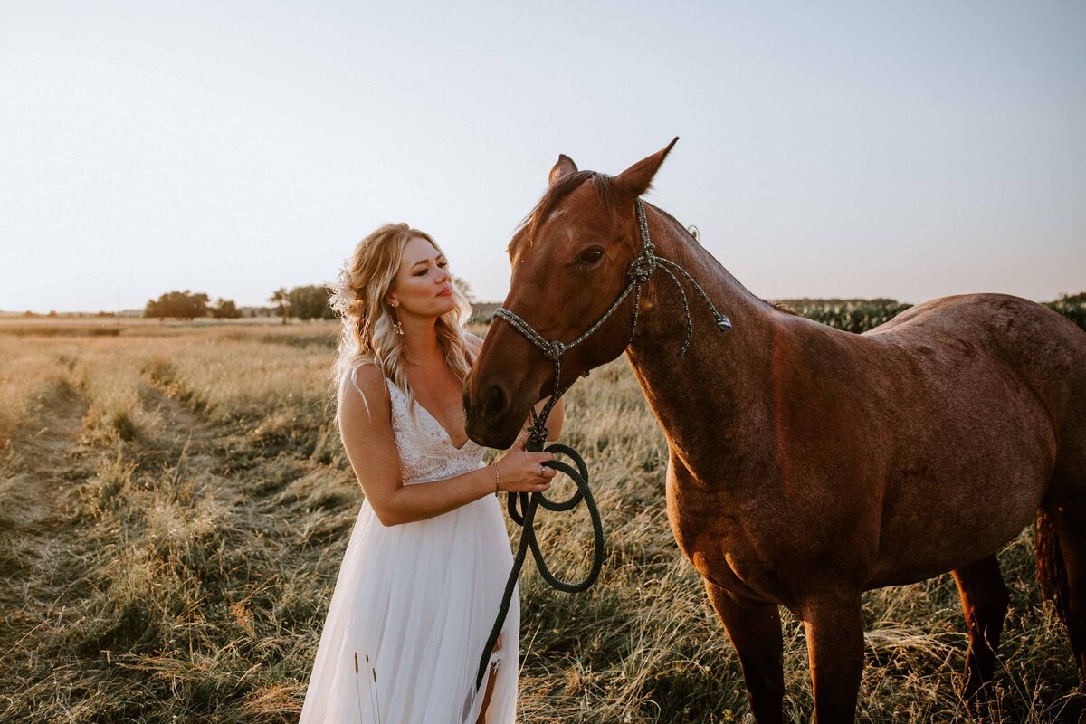 Bride standing at golden hour for wedding day portrait on Exeter, Ontario farm. She is holding her horse's reins and smiling at the brown horse.