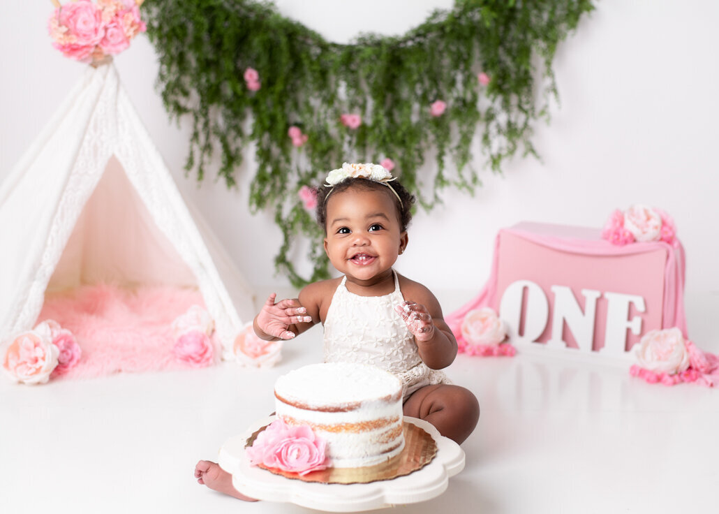 Cake Smash photography -- Smiling baby girl in a white romper enjoys a pink and white cake smash for her first birthday, with floral teepee and greenery backdrop in a Birmingham photography studio