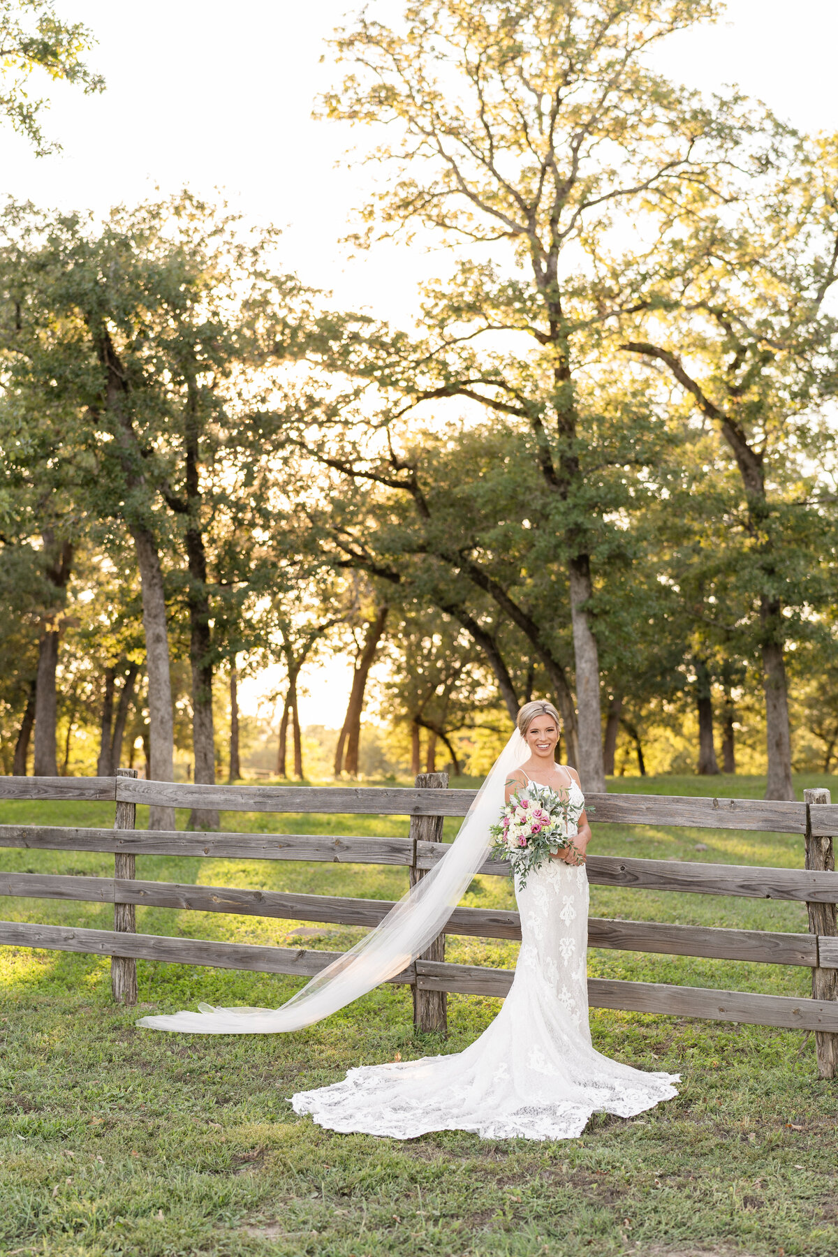 Bride holding bouquet with cathedral length veil in front of an old wooden fence and beautiful tall trees at sunset