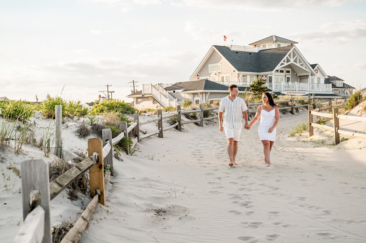 A couple walks hand in hand on a sandy path near beach houses and dunes under a cloudy sky.