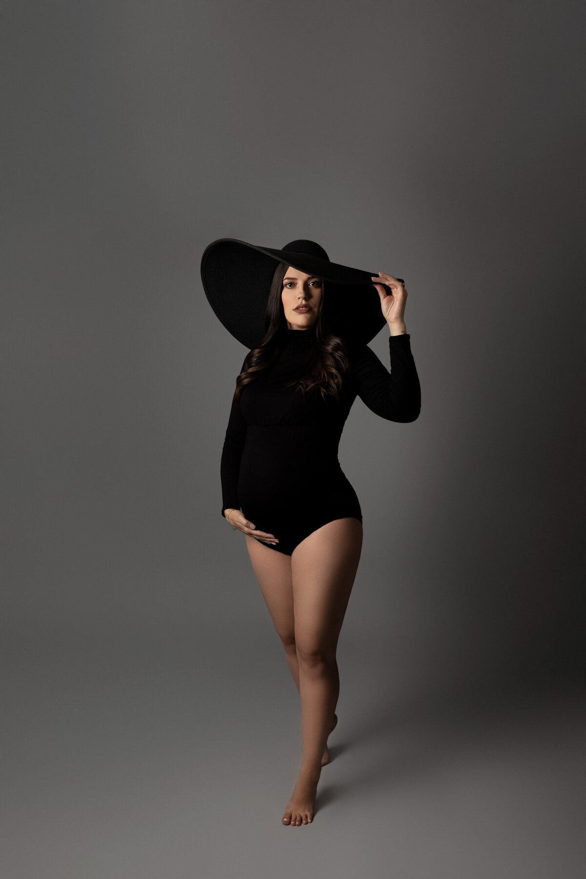 Studio maternity photoshoot in London, Ontario with an editorial vibe. Mom is wearing a black bodysuit and a black, wide-brimmed hat. One hand is resting under her bump, the other hand is touching her hat. She is gently tiptoeing toward the camera. Lots of negative space above mom.