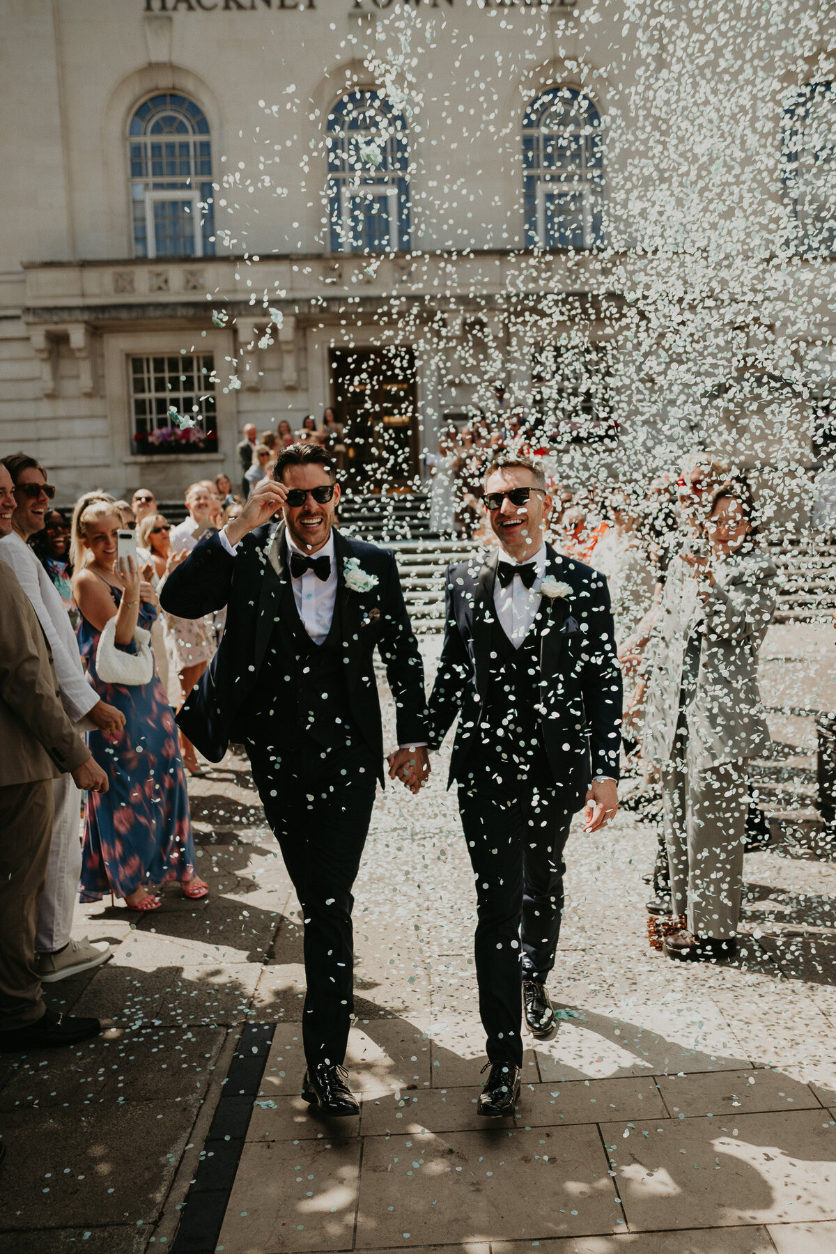 Two grooms walk down the stairs of Hackney Town Hall and through their guests throwing confetti. 