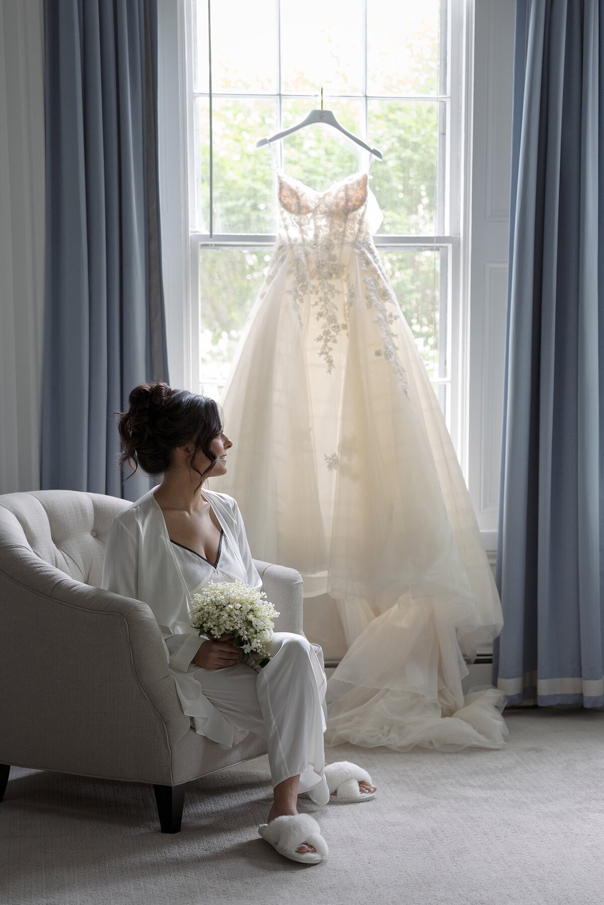 Bride sits near window where her wedding dress is hanging. 