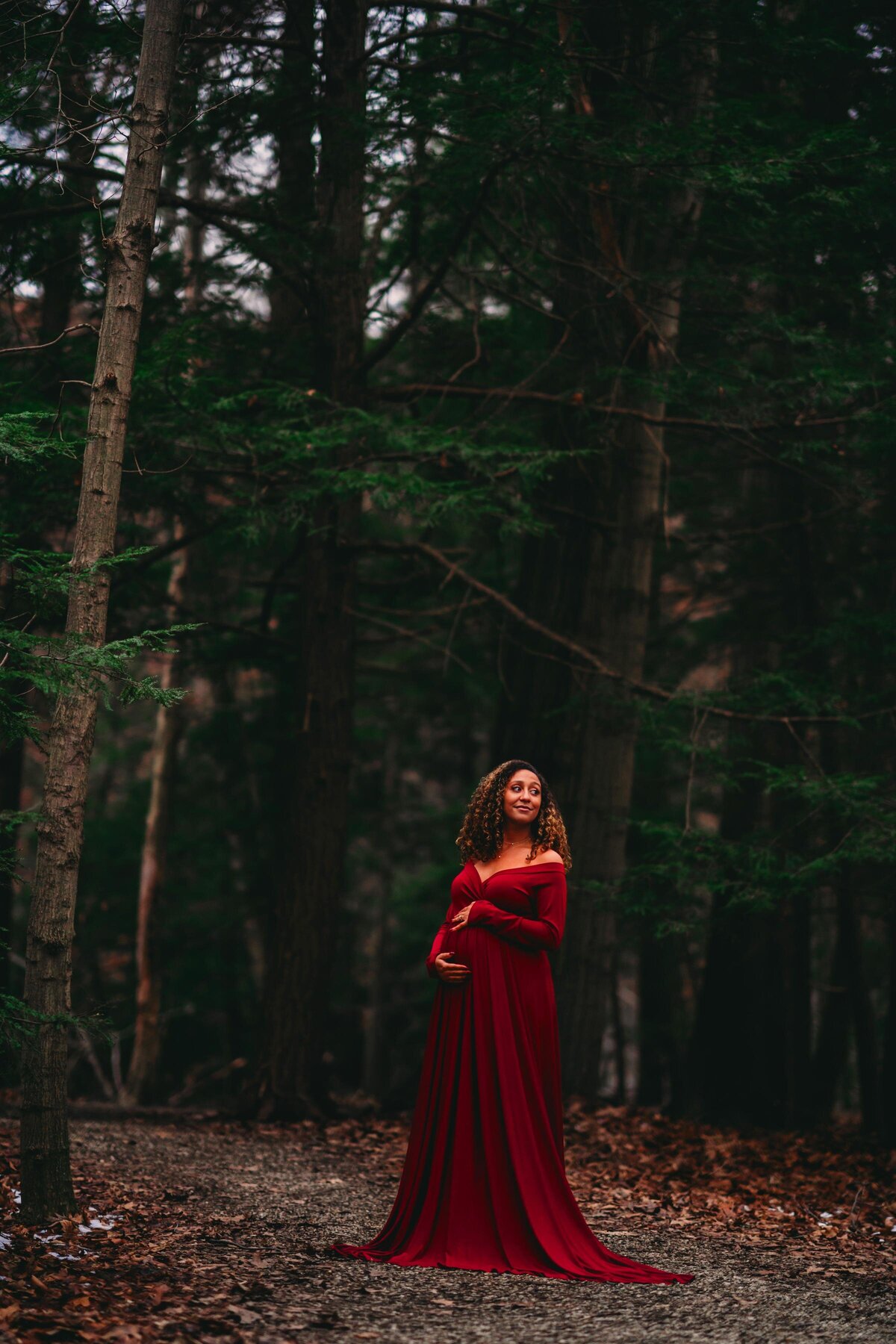 A glowing mother-to-be in a flowing red gown standing in a lush forest, captured in a moody & editorial maternity photography style.