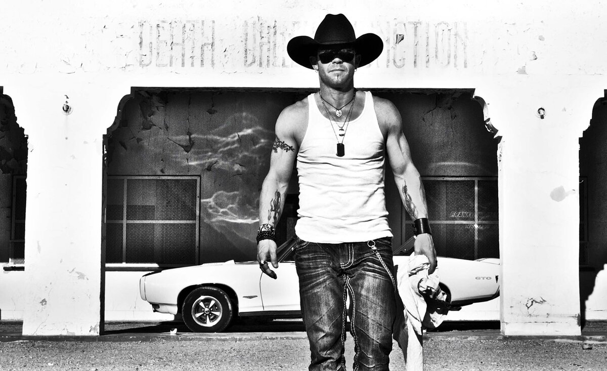 Country music photo Aaron Pritchett black and white wearing black cowboy hat white tank top holding shirt with old convertible behind