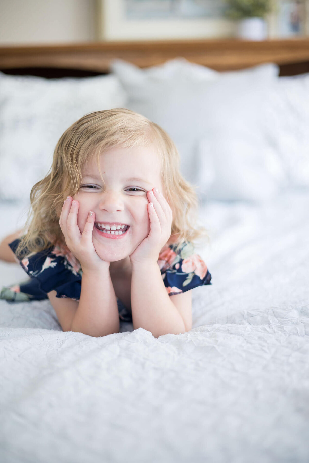 laughing toddler girl in a blue dress with her head in her hands on a bed