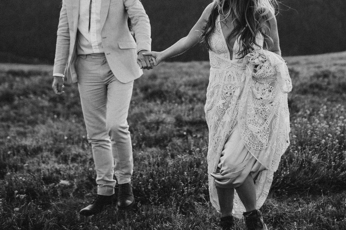 Black and white image of bride and groom wearing a suit and wedding gown holding hands in a field.