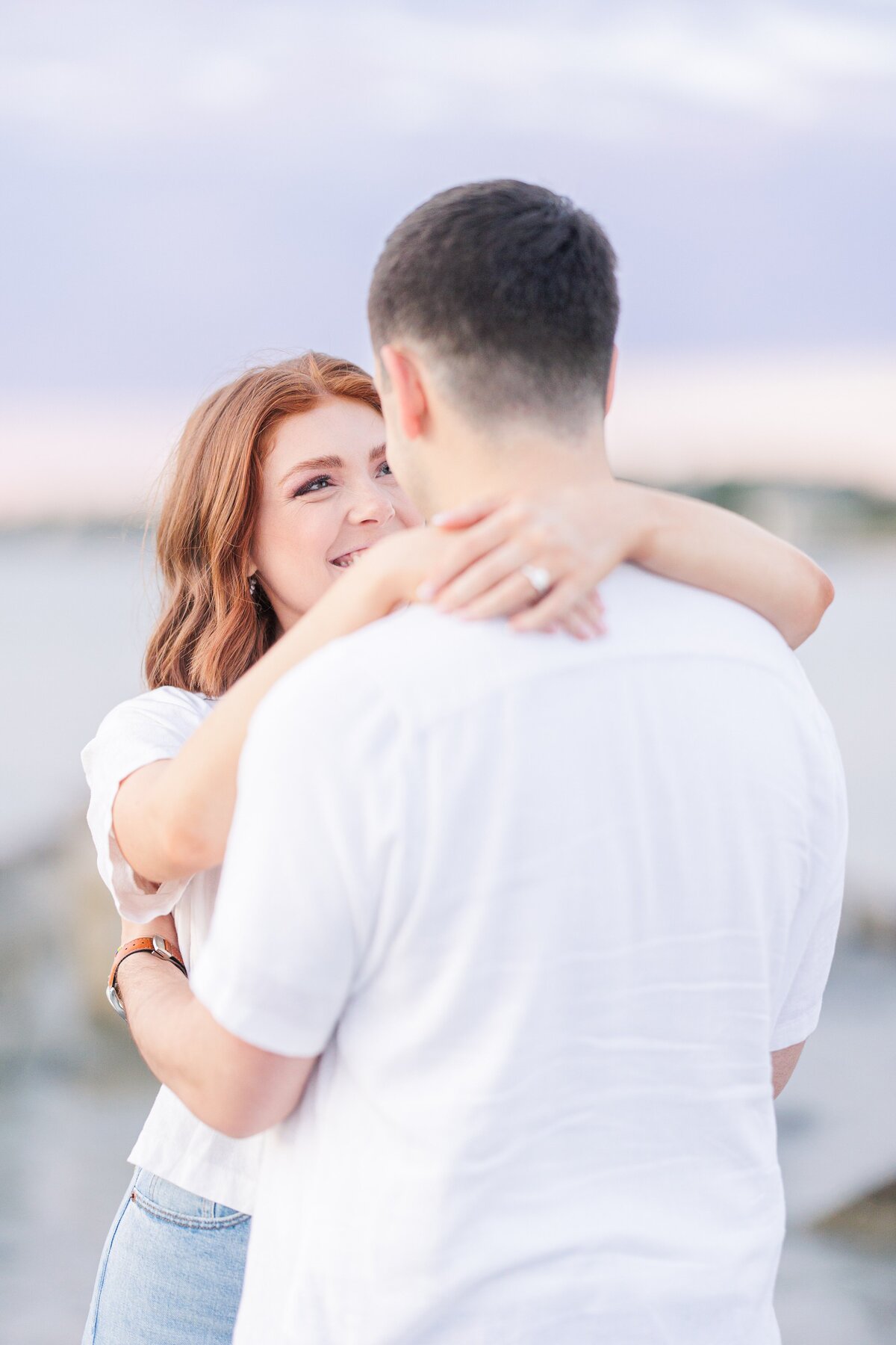 A close up of a man's back as he hugs his fiance and she smiles at him during her Wingaersheek Beach engagement photos.