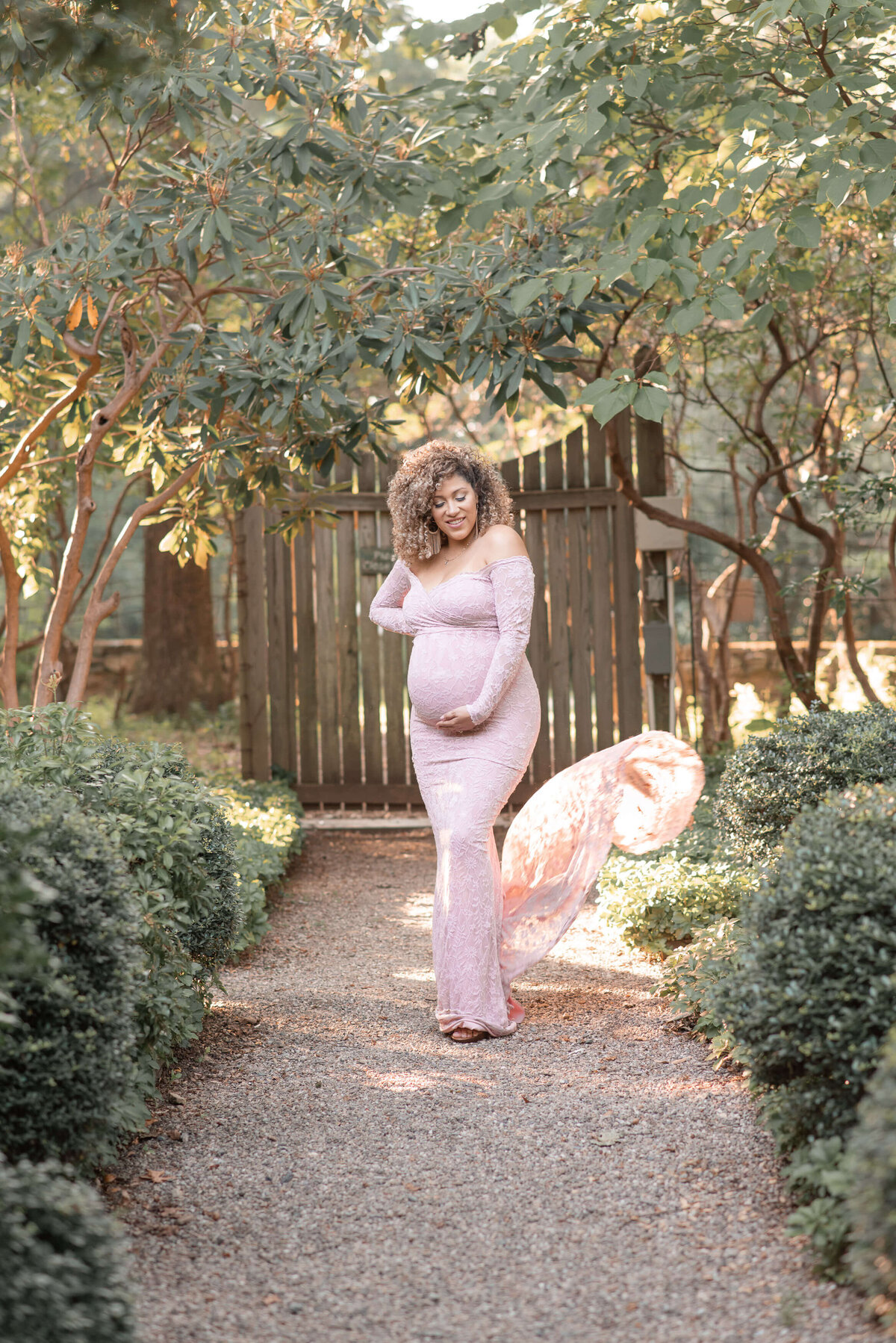 curly hair mom to be wearing a blush maternity dress standing in a garden for her maternity photos by elizabeth klusmann photography