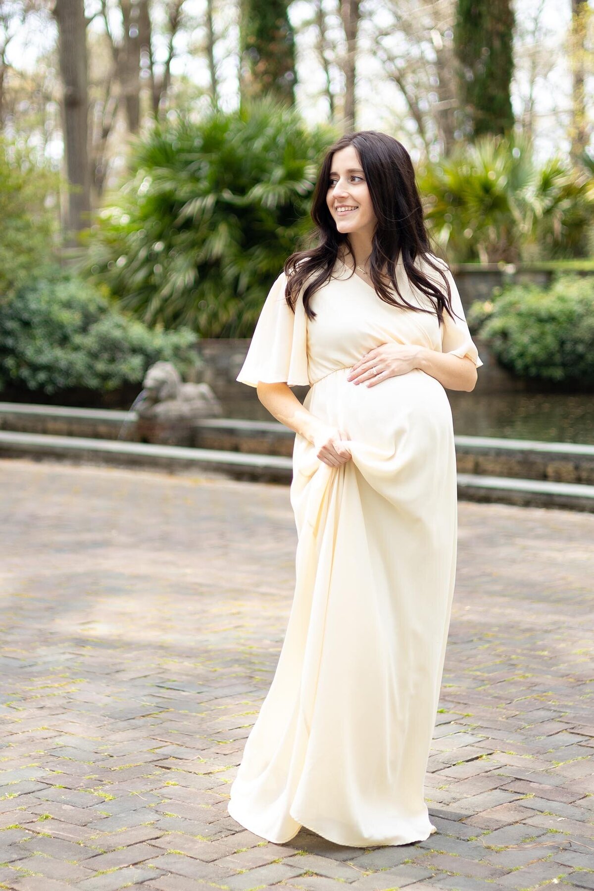A pregnant woman in a cream dress smiles over her shoulder while walking through the Norfolk Botanical Gardens by Virginia Beach Maternity Photographer Mary Eleanor Photography