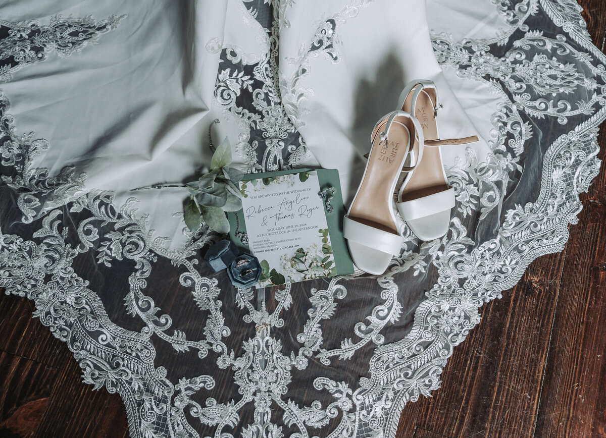 A close-up of beautiful wedding details including elegant bridal shoes, sparkling wedding rings, and a custom invitation suite, artfully arranged on a bride's lace wedding dress at Sweeney Barn in Manassas, Virginia, captured by Quinn Photography LLC.
