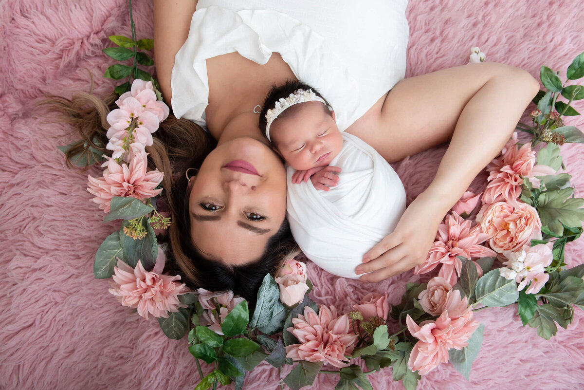 mom laying on the ground with her newborn daughter wrapped in pink in her arms with pink florals for her newborn photography session in Burlington