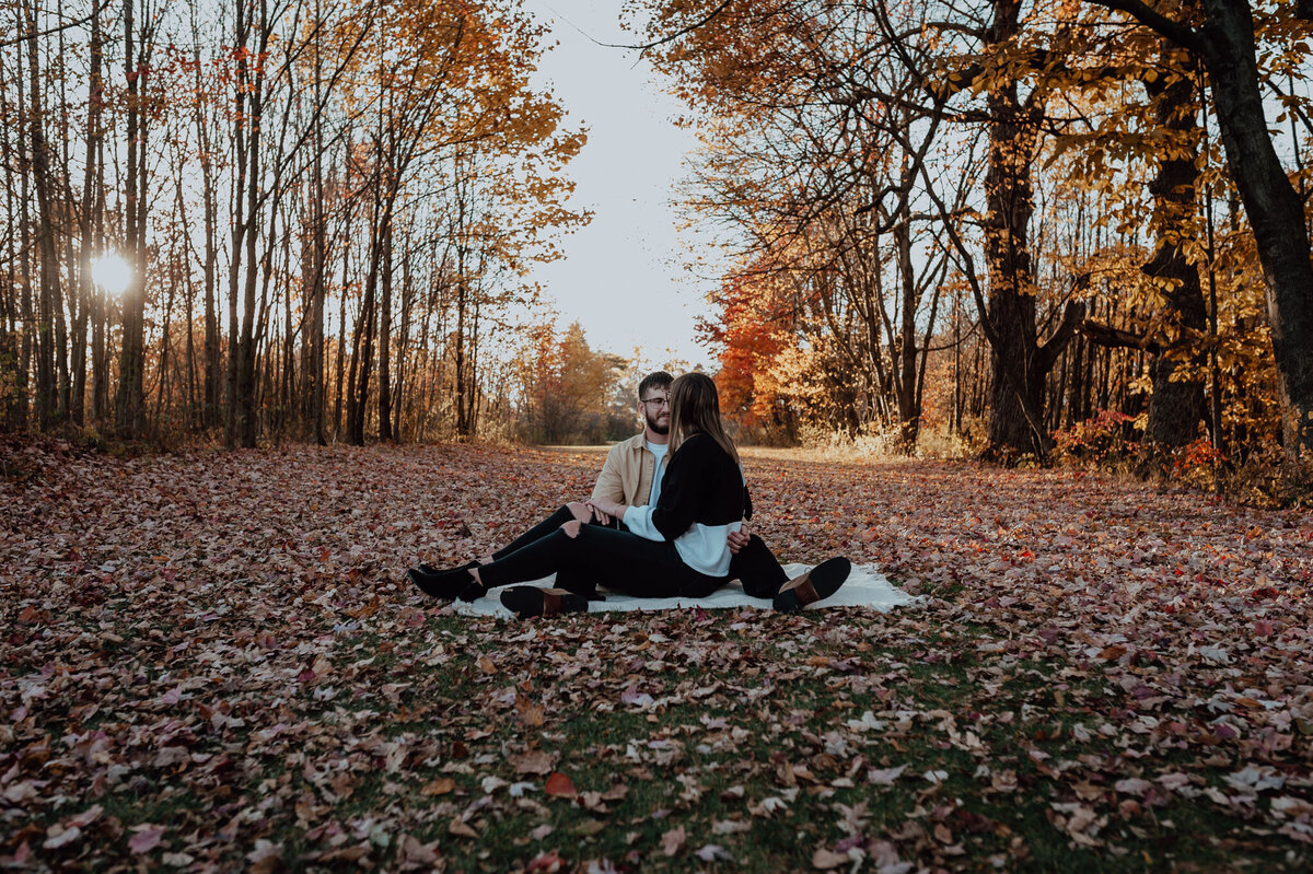 Verona B Photography captures a couple gazing at each other while sitting on a blanket surrounded by crunchy, colorful fall leaves while the sun peaks through the bare trees at Silver Creek Metro Park in Norton, OH.