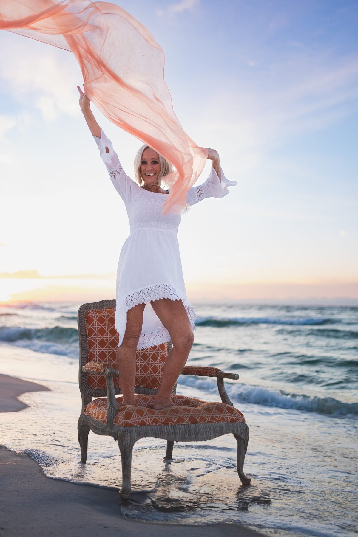 Older woman with white hair wearing a white long-sleeve dress stands on an antique chair on a Florida beach at sunset while waving a peach-colored scarf overhead.