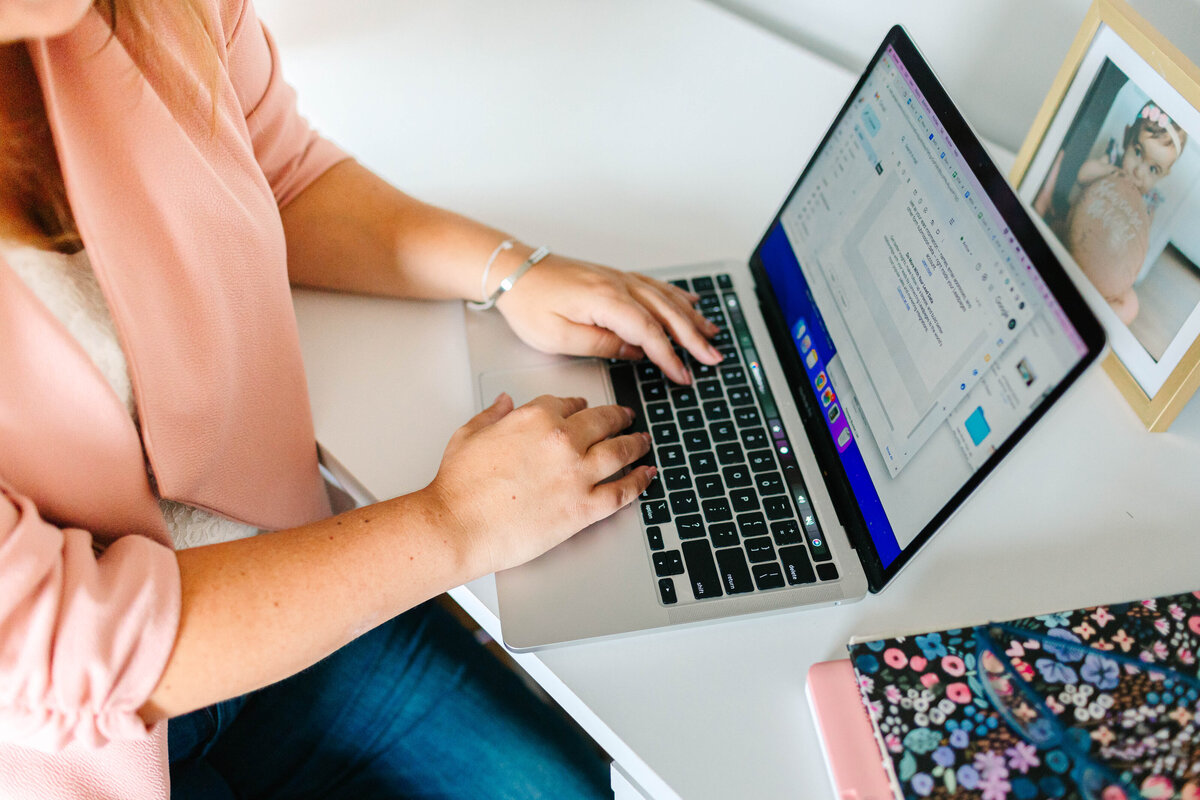 A woman wearing a pink blazer types on a laptop at a white desk. A framed photo of a child, a floral-patterned notebook, and other office essentials are nearby, creating a cozy and organized workspace