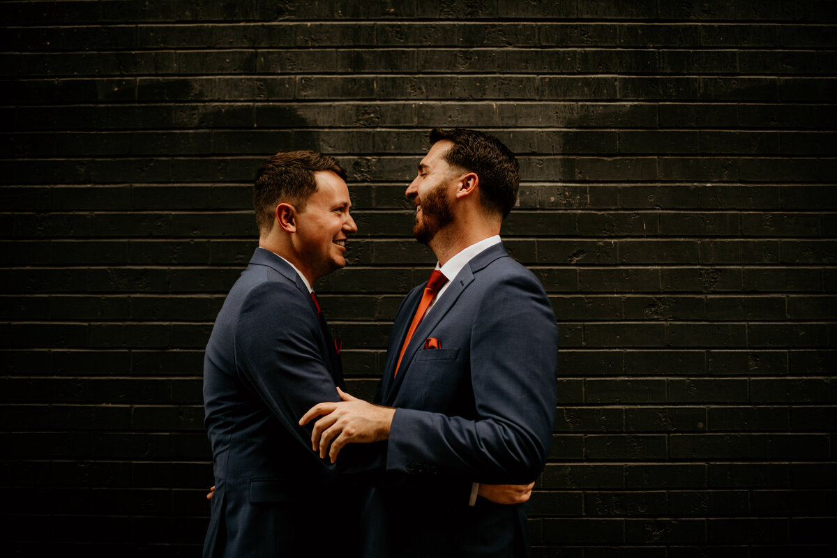 two grooms in blue suits hodling each other in front of a black brick wall in Albuquerque