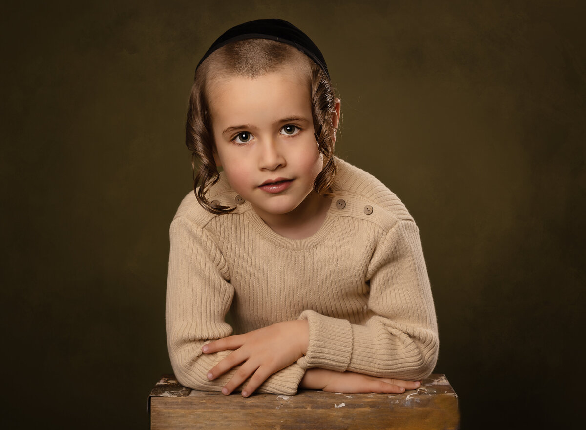 Young boy's portrait. Boy is in a brooklyn ny photo studio with his arms folded on a table in front of him. He is wearing a kippah and looking intently at the camera. Captured by brooklyn family photographer Chaya Bornstein.