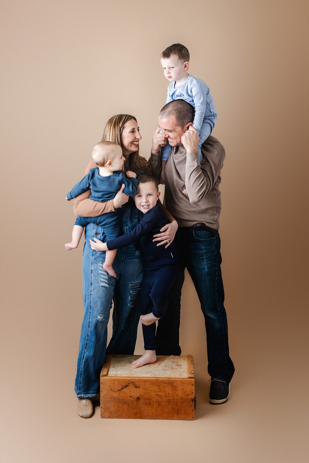 A family of five poses against a neutral background. A woman holds a baby, while a man carrying a child on his shoulders embraces another standing on a wooden box. Captured by a Bloomington, Illinois portrait photographer, everyone is dressed casually and smiling brightly.