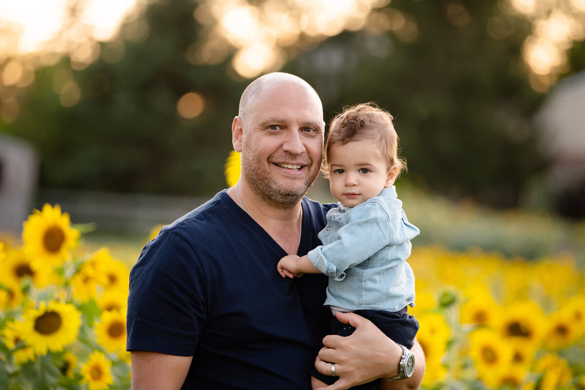 Dad holding his son in the golden hour in front of a field of sunflowers for the summer family photographer session in Burlington, On