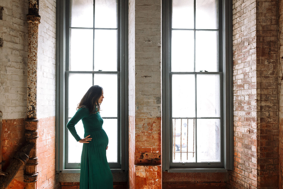 profile of pregnant woman in green dress standing in one of two windows by brick walls at Fort Point in San Francisco