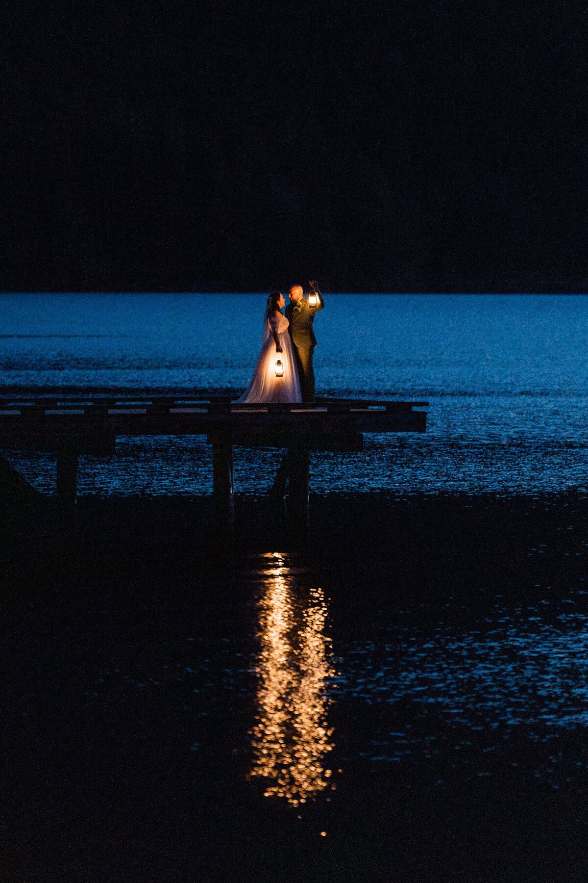 Wide shot of a bride and groom holding lanterns on a pier on Lake Crescent after the sun set. The lanterns are glowing in the water.