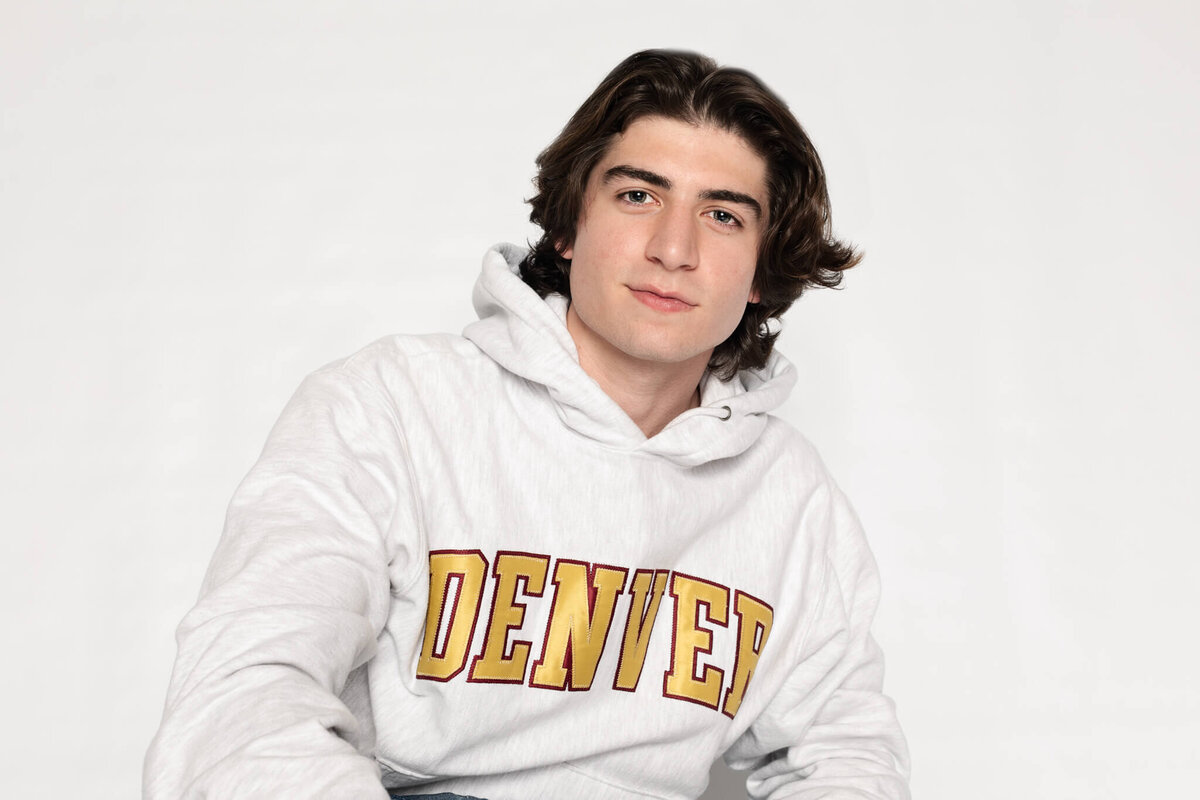 A person with medium-length dark hair is wearing a white hoodie with DENVER written in yellow and red letters. They are sitting against a plain white background, looking directly at the camera with a neutral expression.