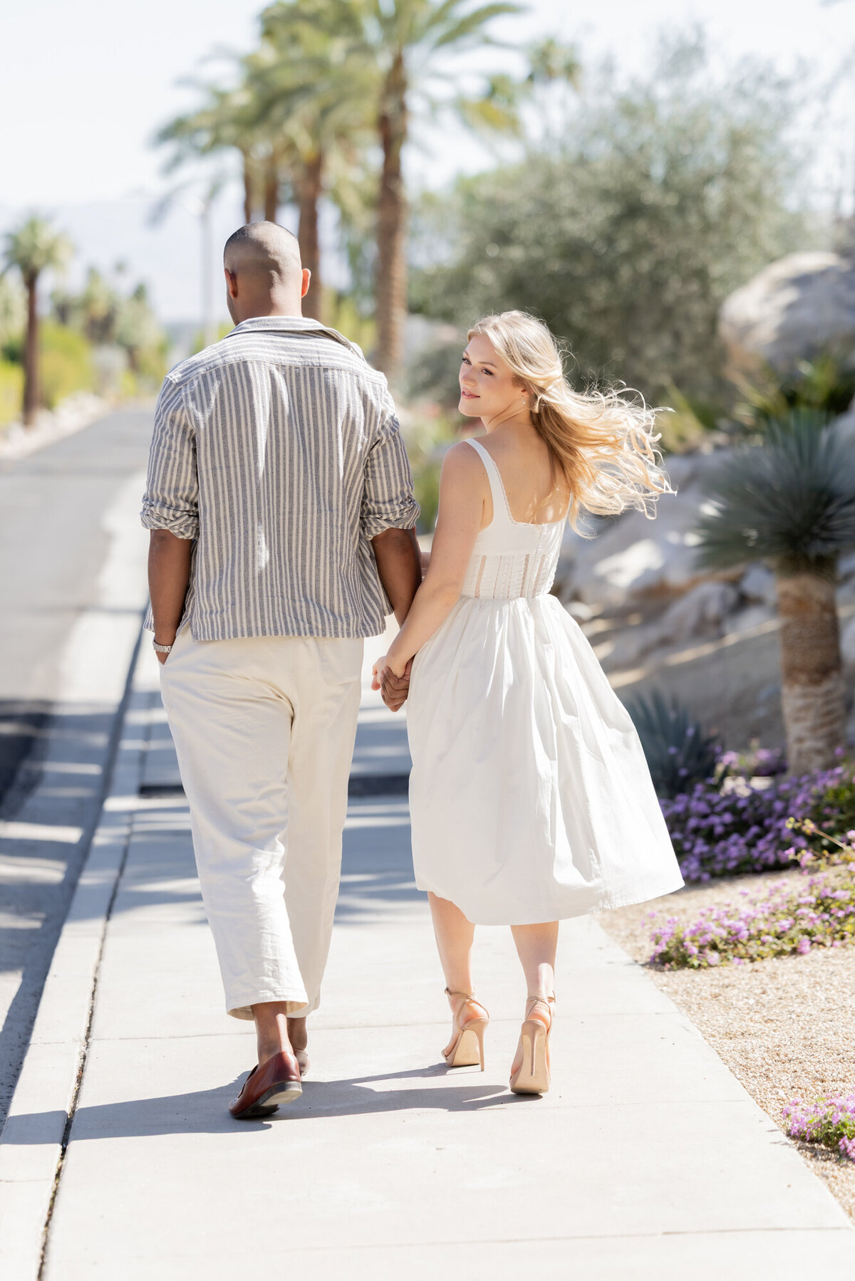 Couple walking hand in hand through a desert garden, surrounded by bright palms and sunlit paths.