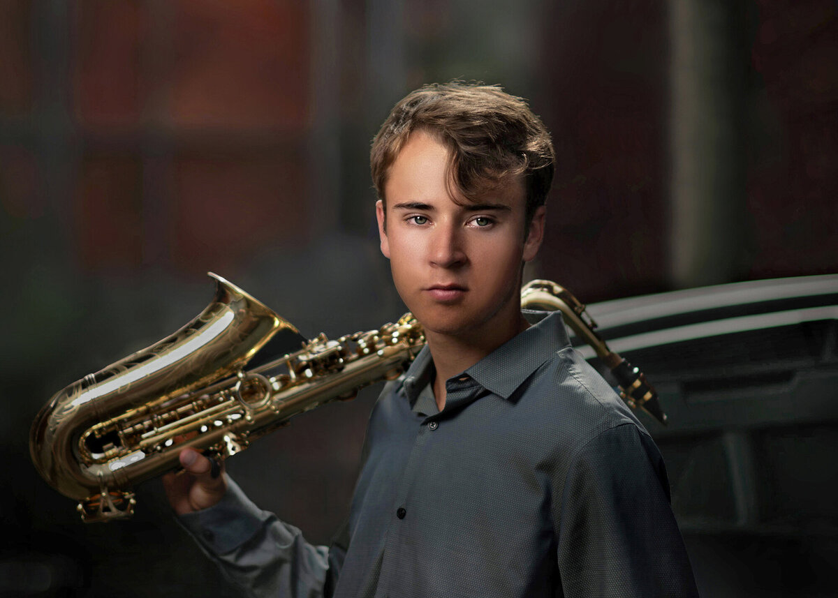 A high school grad in a grey shirt stands in an alley with a saxophone on his shoulder