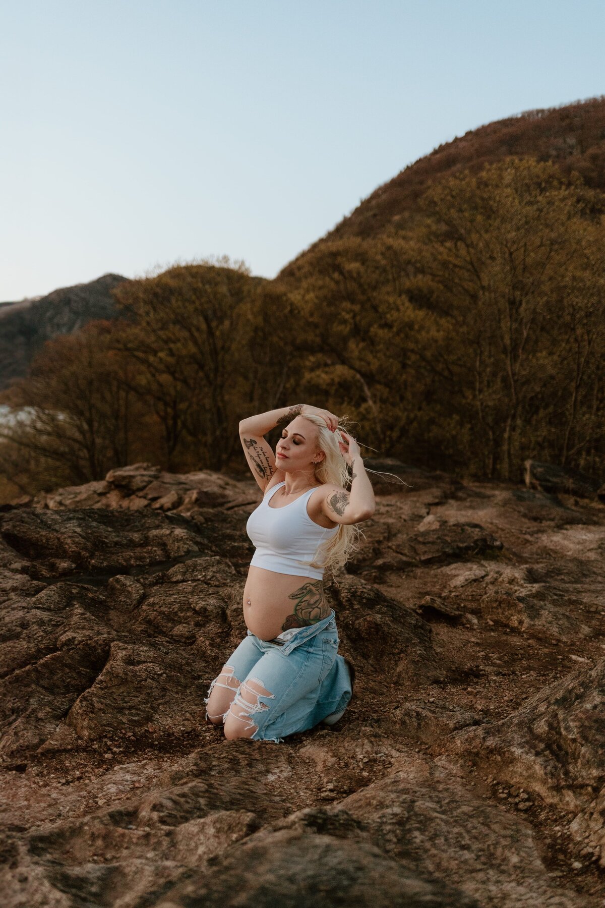 Pregnant woman with blonde hair and tattoos posing confidently on rocky terrain at Little Stony Point, Cold Spring. She is wearing a white tank top and distressed jeans, with her hands gently touching her hair as she enjoys the serene natural backdrop.