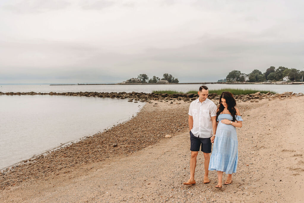 Expecting couple walking together along Connecticut shoreline in coordinated blue outfits